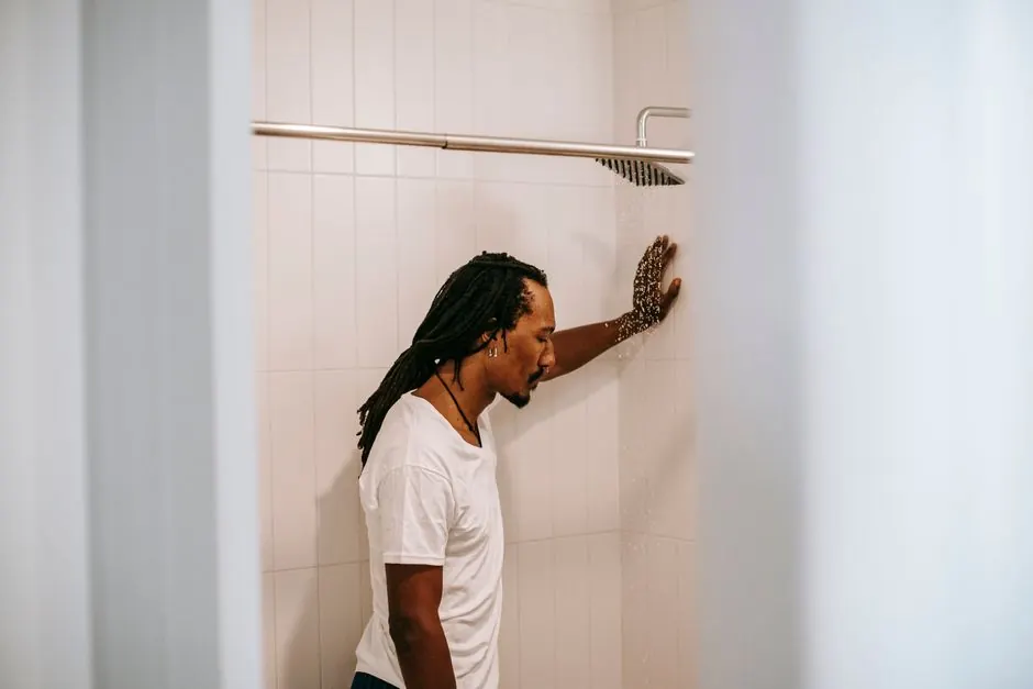 Side view of sad young informal black guy with Afro braids in white t shirt standing in shower booth under water stream and leaning on wall